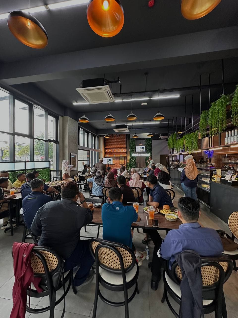 Guests sharing a long table inside The Komunal café, brick wall and greenery behind them