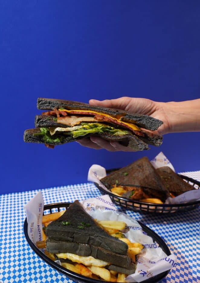 A hand holding a charcoal club sandwich above a basket of fries